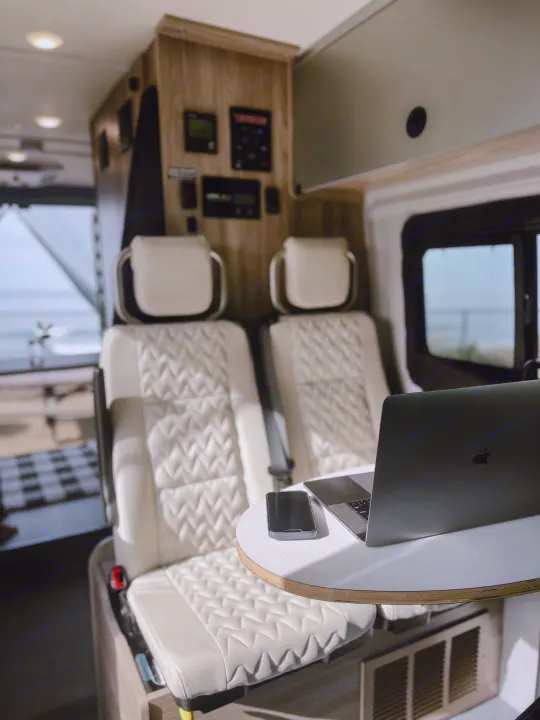 Comfortable RV rental interior with light-colored, quilted seats, a laptop, and phone on a small table. Ocean view outside.. Winnebago Solis 59PX 2023