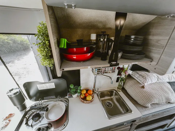 Cozy RV rental kitchen featuring a compact stove, sink, organized shelves with dishes & cookware, and a comfy sleeping area.. Winnebago Solis 59PX 2023