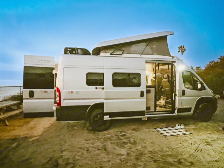 This Winnebago Solis rv rental, with open doors and pop-top, is parked on sand near the ocean, ready for a cozy camp.. Winnebago Solis 59PX 2023