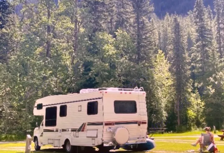 A white RV rental is parked at a scenic forest campsite. Enjoy outdoor living with towering trees and a person relaxing nearby.. Ford Econoline 1988