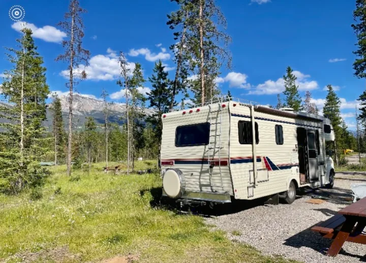 Cream RV rental at a sunny mountain campsite with pine trees, grassy areas, and distant peaks.. Ford Econoline 1988