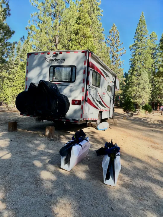 An rv rental is parked at a pine tree campsite, with bicycles on the back and two Oru Kayaks on the ground.. Coachmam Freelander 21QB 2012