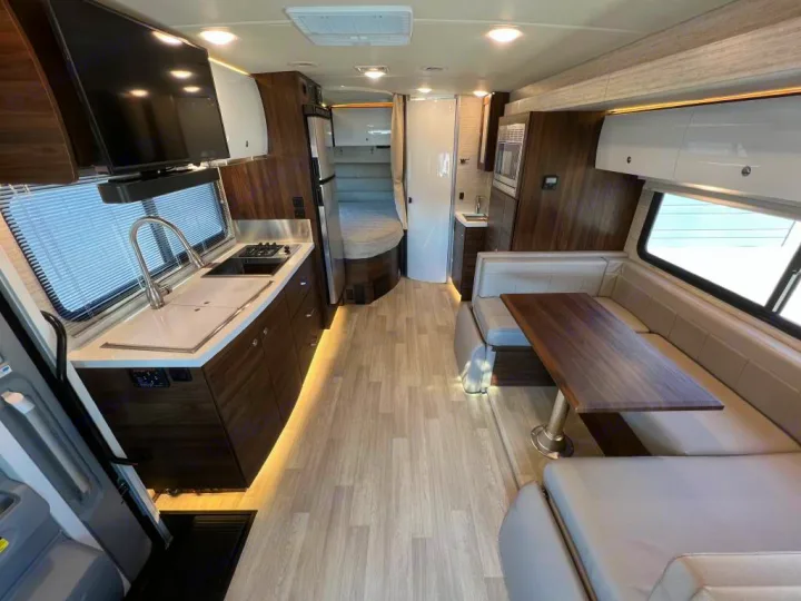 Modern RV rental interior featuring kitchen, dining, and a rear bedroom. Light wood floor & warm lighting enhance comfort.. Winnebago View 2023