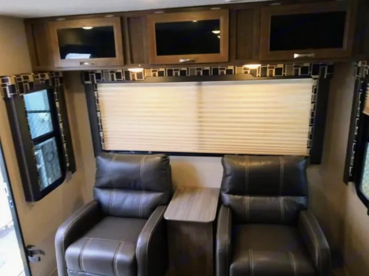 View inside an RV rental showing two dark recliners, an end table, large window with blinds, and overhead cabinets.. Dutchmen Coleman Light 2018