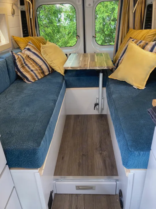 Cozy RV rental dinette with comfortable blue seating, bright pillows, and a compact wooden table, ready for travel adventures.. Ram 3500 Extended 2021