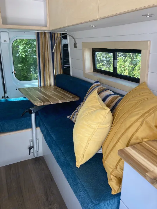 This inviting RV rental camper van interior features blue upholstered seating, a wood table, and sunlit windows with tree views.. Ram 3500 Extended 2021