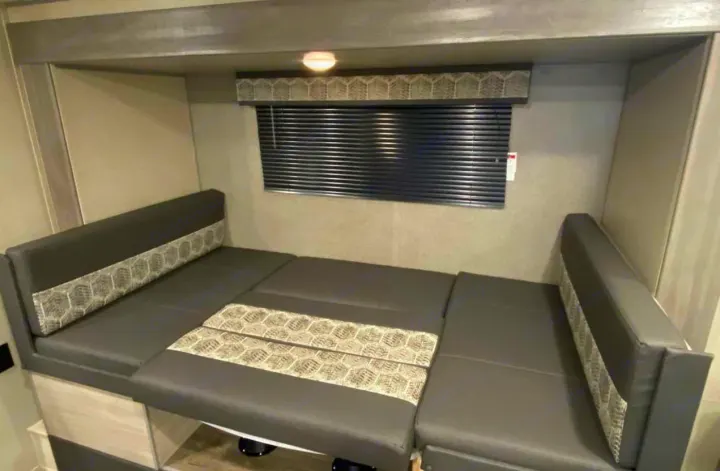 Compact RV rental dinette converts to a bed. Grey seating with patterned fabric, window blinds, and overhead light in a modern interior.. Forest River Coachmen Catalina Summit 2022