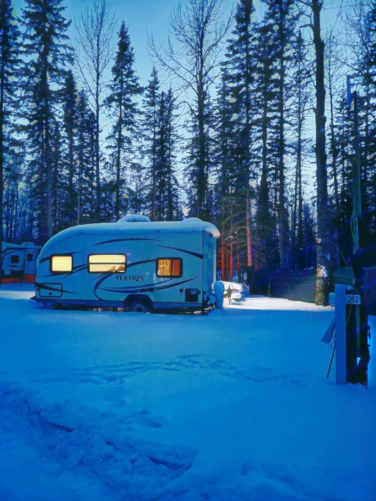 Cozy winter RV rental in a snowy forest at night. Warm lights glow from the camper, surrounded by tall trees.. Gulf Stream Other 2018
