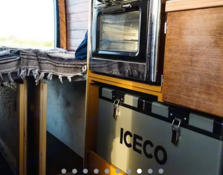 Compact RV rental interior shows a cozy bed with blanket, a mini oven, and an ICECO fridge/freezer unit. Perfect for road trips!. Mercedes Sprinter 2013