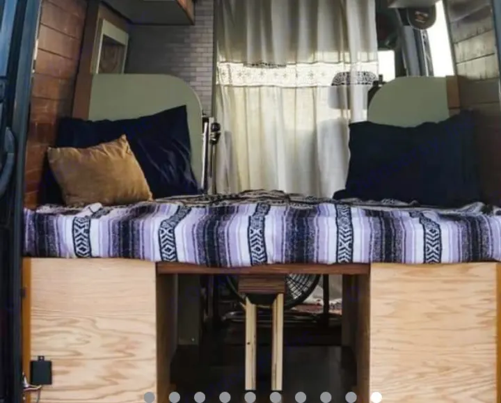 RV rental van interior: A cozy bed with a striped blanket & pillows, flanked by wood walls. Under-bed storage visible. White curtains in back.. Mercedes Sprinter 2013