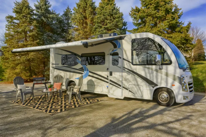 Relaxing by your RV rental! This white motorhome with an extended awning, outdoor chairs, and grill is perfect for camping adventures.. Thor Vegas 2022