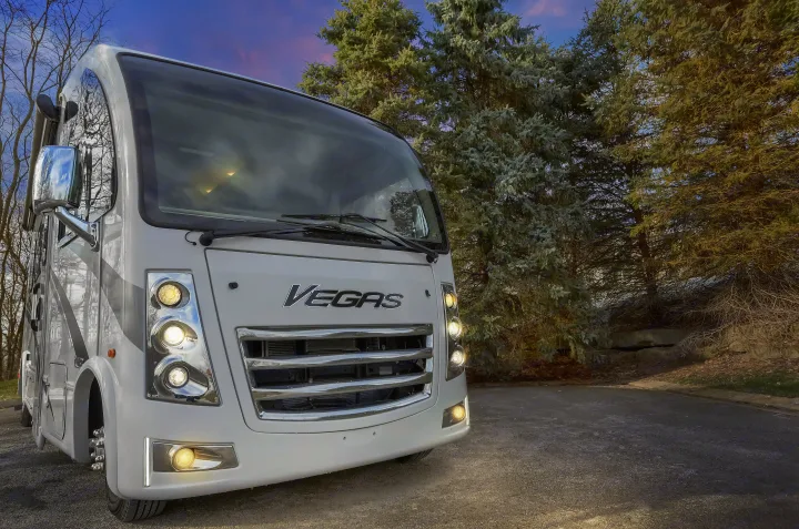 Front view of a modern white 'Vegas' RV, its bright lights on, parked on pavement at dusk. Ideal for rv rental adventures.. Thor Vegas 2022
