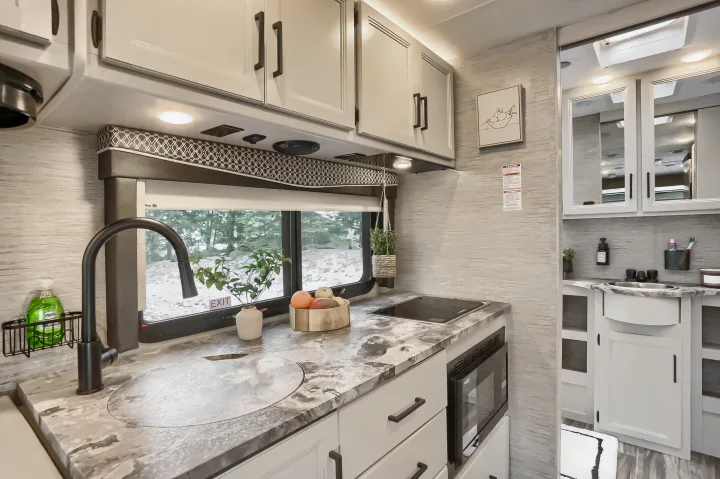 Bright RV rental kitchen with light cabinetry, sink, cooktop, and window view. Modern interior leads to a compact bathroom.. Thor Vegas 2022