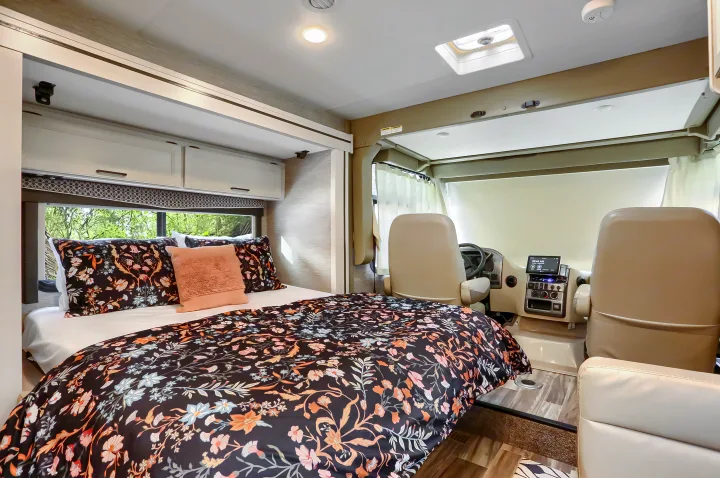 Inside an RV rental: a cozy bedroom with floral bedding seamlessly connects to the front cockpit area with tan seats.. Thor Vegas 2022