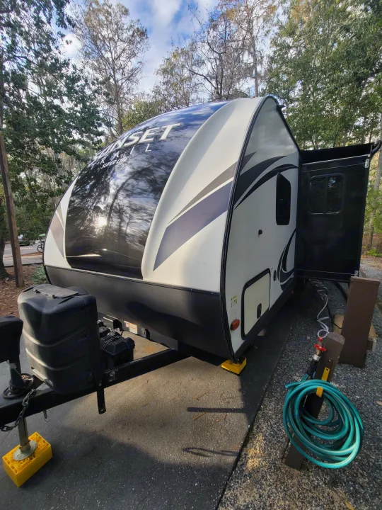 This white and black rv rental travel trailer is parked at a shaded campsite, awning deployed, with water & power hookups.. Crossroads RV Sunset Trail Super Lite 2018