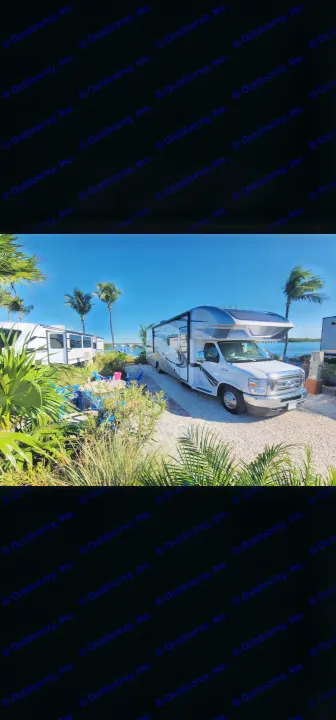 A white RV rental on a gravel site by clear blue water, surrounded by lush palm trees and tropical foliage. Ideal vacation spot.. Entegra Esteem 29v 2022