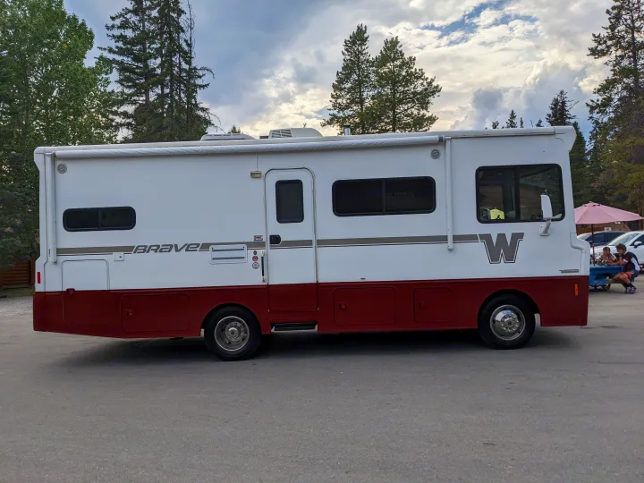 A white and red Brave rv rental, seen from the side, is parked on asphalt with trees & cloudy sky.. Winnebago Brave 2015