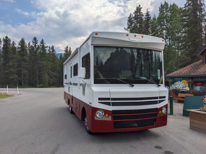 Class A Winnebago rv rental parked on asphalt, surrounded by tall trees & mountains reflected in its windshield.. Winnebago Brave 2015