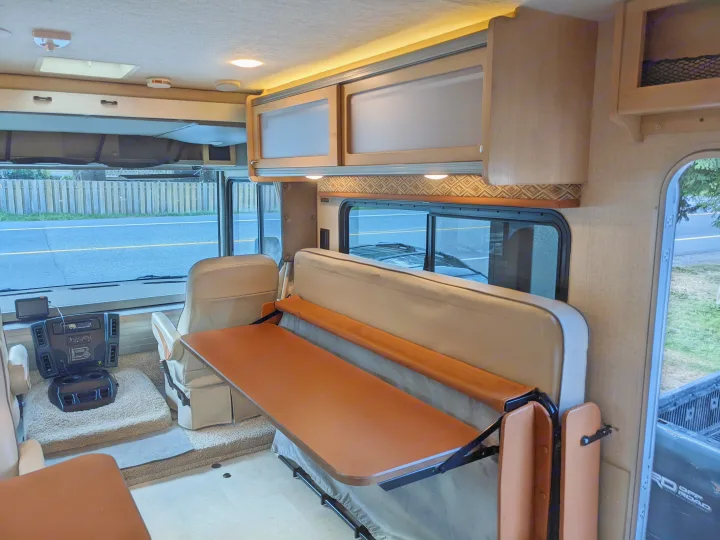 Inside an RV rental: front cockpit with two seats, a fold-down table in the dinette area, and overhead storage cabinets.. Winnebago Brave 2015