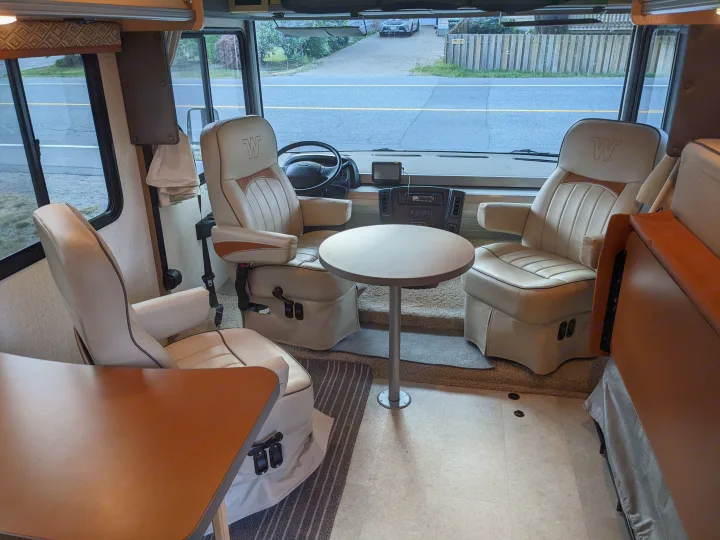 Inside an RV rental: driver and passenger captain's chairs, a small table, and the cockpit dash, offering a view outside.. Winnebago Brave 2015