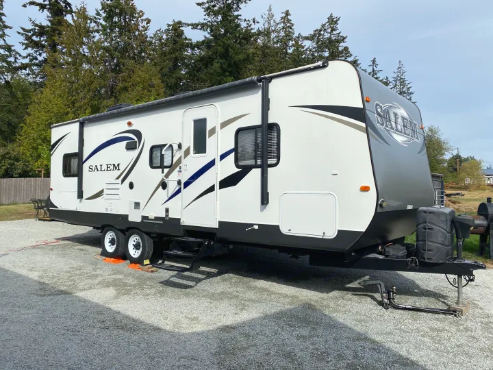 A large, modern white and grey Salem travel trailer for rv rental, parked on a gravel lot with trees in the background.. Forest River Salem 2015
