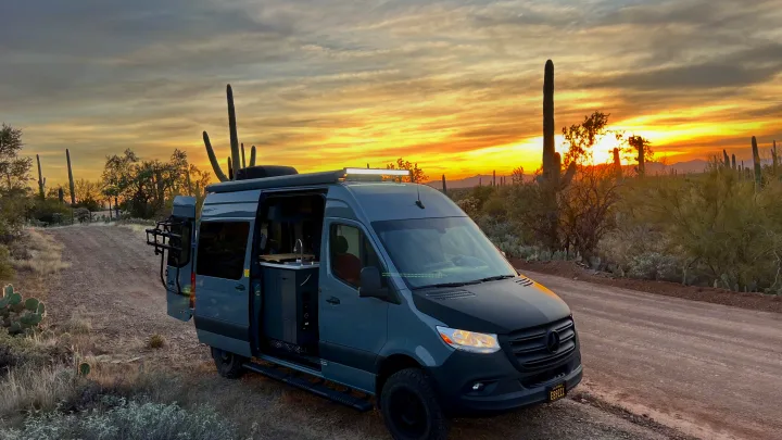 Gray Mercedes Sprinter rv rental on a dirt road, door open, showcasing its interior. Desert sunset, saguaro cacti.. Synergy Mercedes-Benz Sprinter 2022