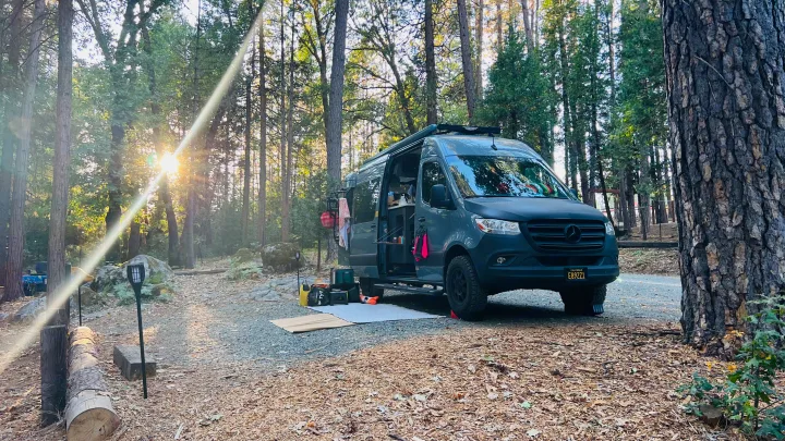 This dark gray RV rental camper van sits in a sunny forest campsite, fully set up for a serene camping adventure.. Synergy Mercedes-Benz Sprinter 2022