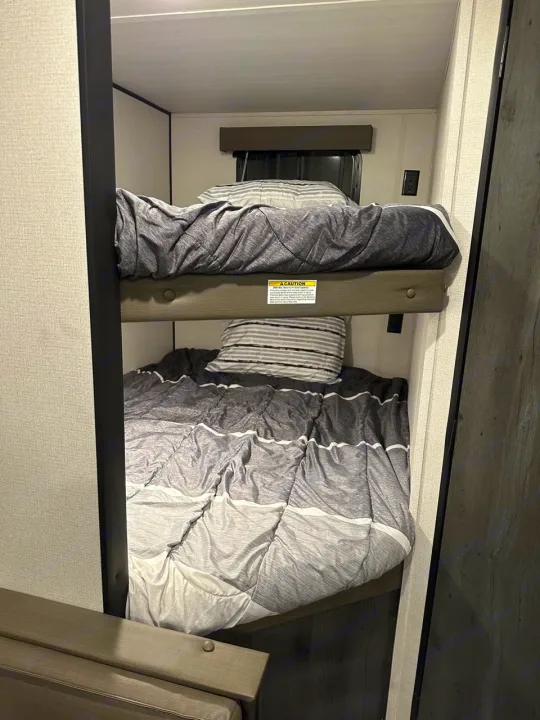 This RV rental features a comfortable bunk bed with gray bedding and accessible power outlets, ideal for travel.. Keystone RV Hideout 2022