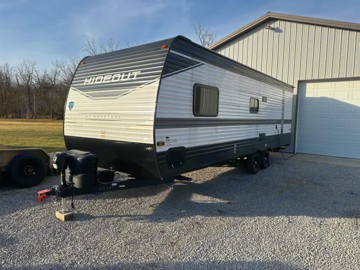 A gray & black Keystone Hideout travel trailer, ideal for RV rental, parked on gravel beside a white barn under a clear sky.. Keystone RV Hideout 2022