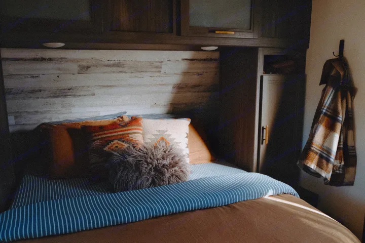 Cozy RV rental bedroom bathed in warm sunlight. Features a wood headboard, patterned pillows, and a hanging plaid shirt.. Dutchmen Kodiak Cub 2020