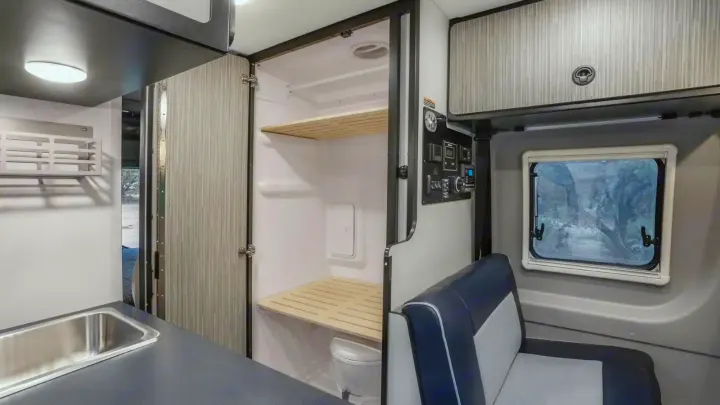 Inside an RV rental: features a kitchen sink, compact wet bath with toilet & wooden shelves, comfy bench seat, and control panel.. Winnebago Revel 2023