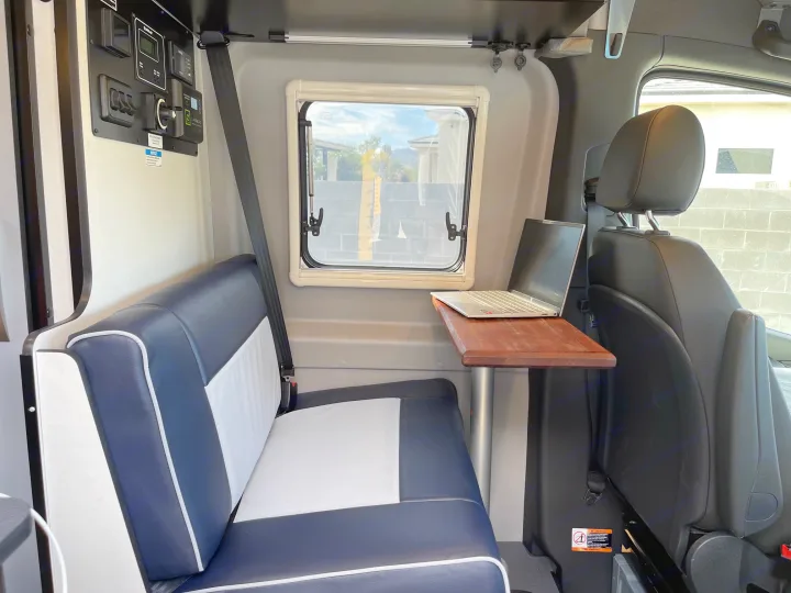 Compact rv rental interior showing blue & white seating, a window, control panel, and a laptop on a wooden table.. Winnebago Revel 2023