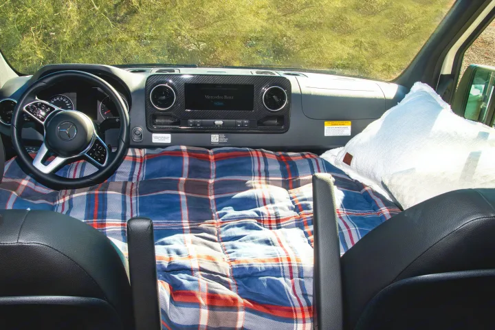 RV rental Mercedes-Benz van interior showing dashboard and a plaid blanket bed setup with pillow.. Winnebago Revel 2023