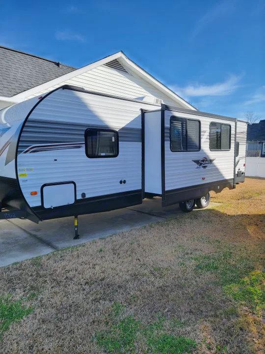 Side view of a modern white and grey travel trailer, ideal for an RV rental, parked on concrete next to a house on a sunny day.. Forest River Shasta 2022