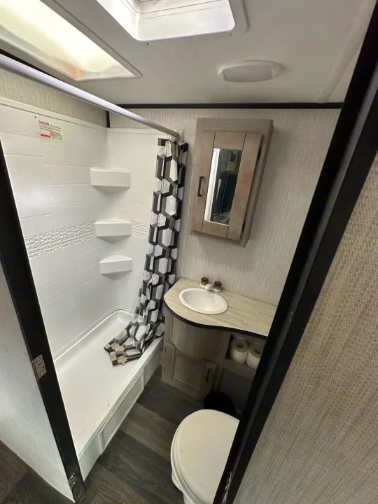 Inside a clean RV rental bathroom: compact shower with patterned curtain, corner shelves, sink vanity, toilet, and medicine cabinet.. Heartland Other 2021