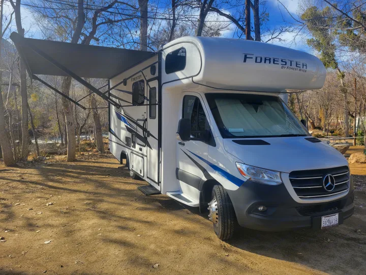 RV rental: A white Forester Class C motorhome with an awning extended, parked by a river at a sunny campground.. Forest River Forester 2023