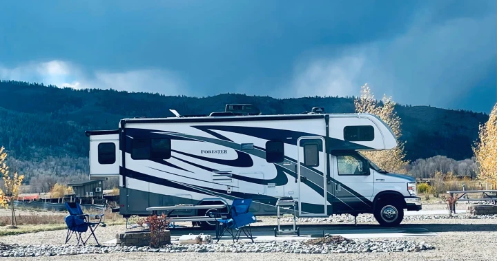 Large white and gray rv rental parked at a gravel campsite with blue chairs, a table, and fire pit. Mountains under a cloudy sky.. Forest River Sunseeker TS 2020