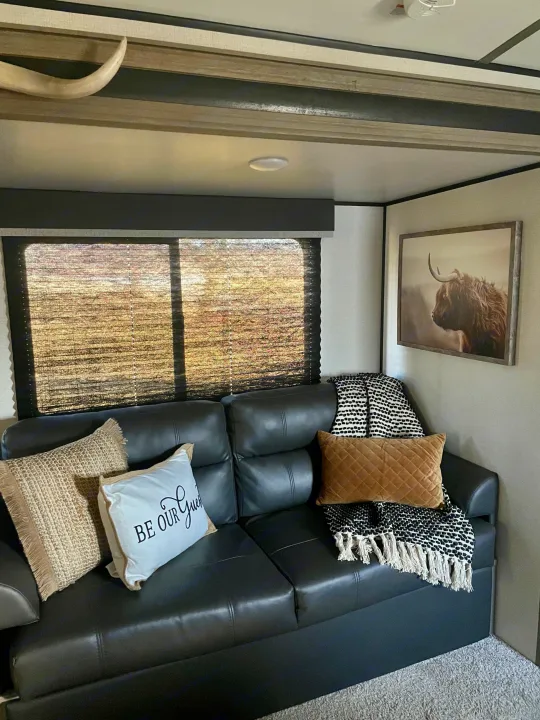 Comfortable RV rental living space featuring a dark couch, decorative pillows, a cozy blanket, and Highland cow art.. Keystone RV Springdale 2021