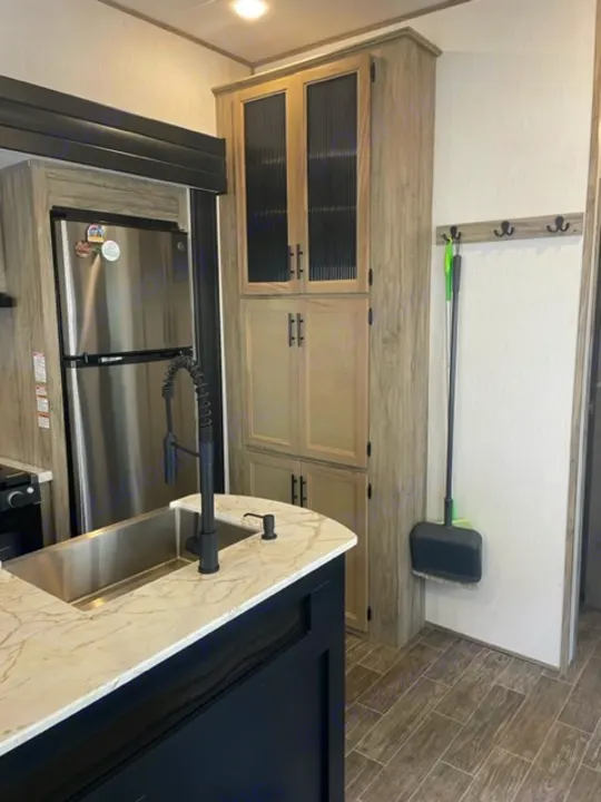 Modern RV rental kitchen with a large sink, stainless steel refrigerator, and wood-toned cabinets. A broom hangs on the wall.. Forest River Cherokee Arctic Wolf 2022