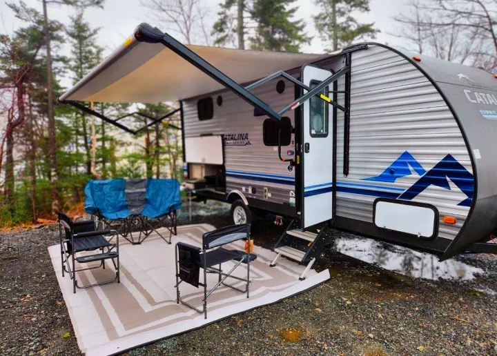 Explore nature! This **RV rental**, a Catalina travel trailer, offers a cozy setup with an awning, rug, and chairs at a wooded campsite.. Forest River Coachmen Catalina Summit 2022