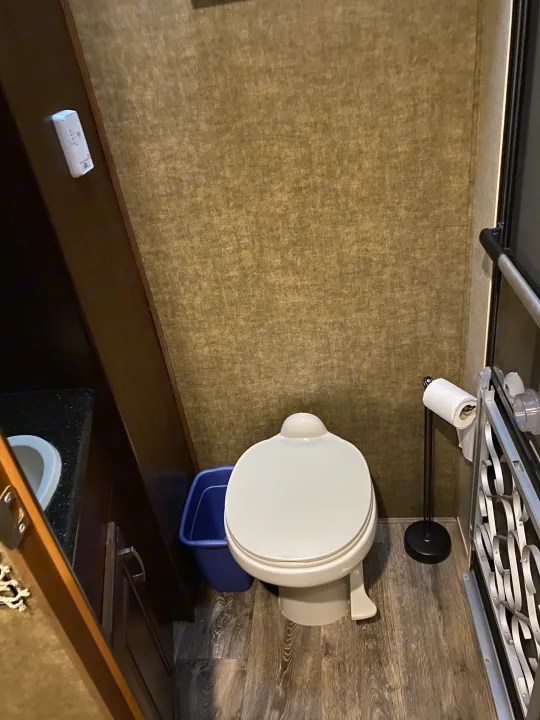 Compact RV rental bathroom: toilet, small sink, blue trash can, and textured walls with wood-look floor.. Heartland RVs Gateway 2015