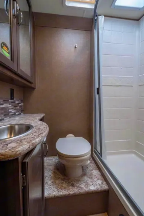 Compact RV rental bathroom featuring a sink, toilet on a raised platform, and a separate shower stall with glass door. Ideal for travel.. Thor Four Winds 2019