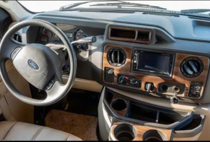 Interior of an rv rental dashboard, showing a Ford steering wheel, infotainment screen, wood grain trim, and lower cup holders.. Thor Four Winds 2019