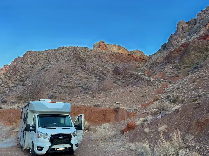 A white RV rental camper van rests on a dirt path, surrounded by arid, rocky mountains and sparse desert plants.. Coachmen Crosstrail 20XG 2022