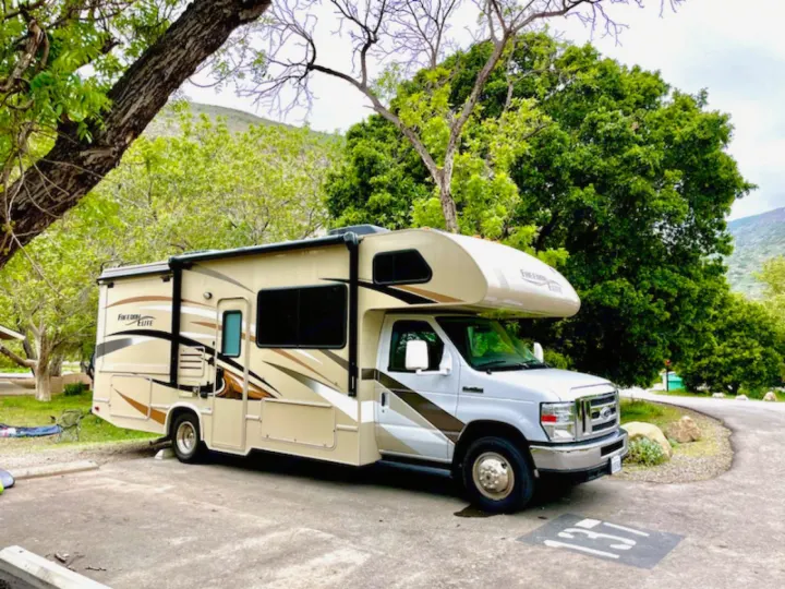 A beige Freedom Elite RV rental sits parked at campsite 137. Lush green trees and hills surround this perfect travel spot.. Thor Freedom Elite 2017