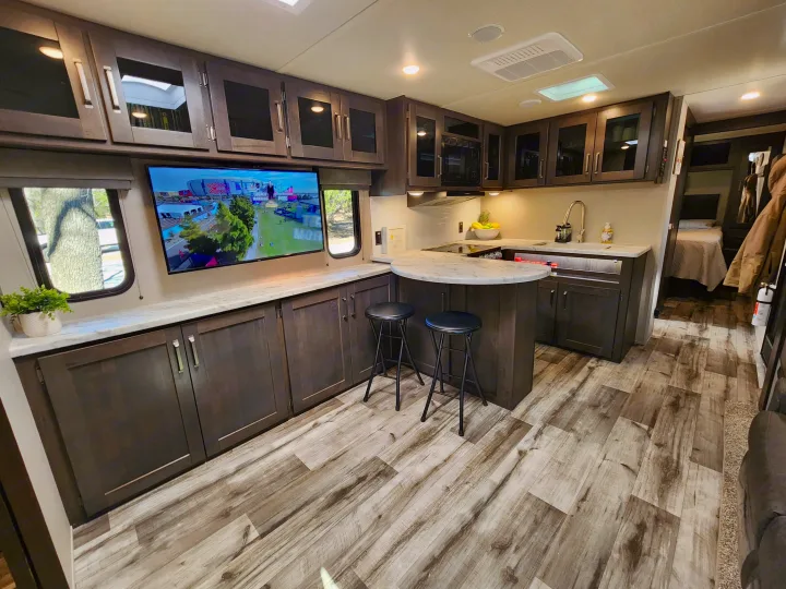 Modern RV rental interior with a spacious kitchen, island seating, and living area featuring a TV and wood-look flooring.. Grand Design Transend Xplor 315BH 2023