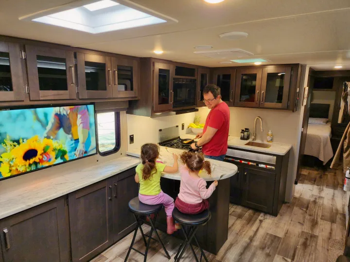 RV rental interior: Dad cooks for two girls in a well-equipped kitchen. Bright and spacious mobile home for family adventures.. Grand Design Transend Xplor 315BH 2023