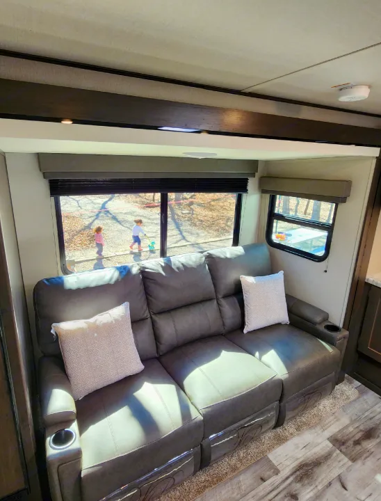 Cozy RV rental interior: large gray sofa with cup holders, natural light, and kids enjoying the outdoors.. Grand Design Transend Xplor 315BH 2023