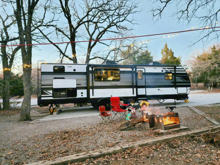 An RV rental is parked at a gravel campsite. Two children are seated by a burning campfire with string lights overhead.. Grand Design Transend Xplor 315BH 2023