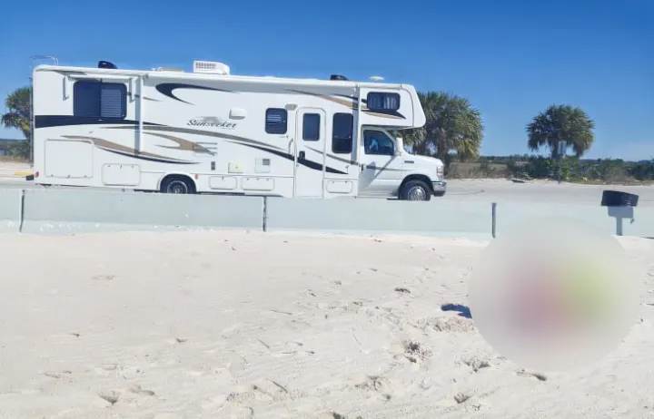 A white RV rental with brown graphics is parked beside a sandy beach, separated by a concrete barrier, under a clear sky.. Forest River Sunseeker 2012